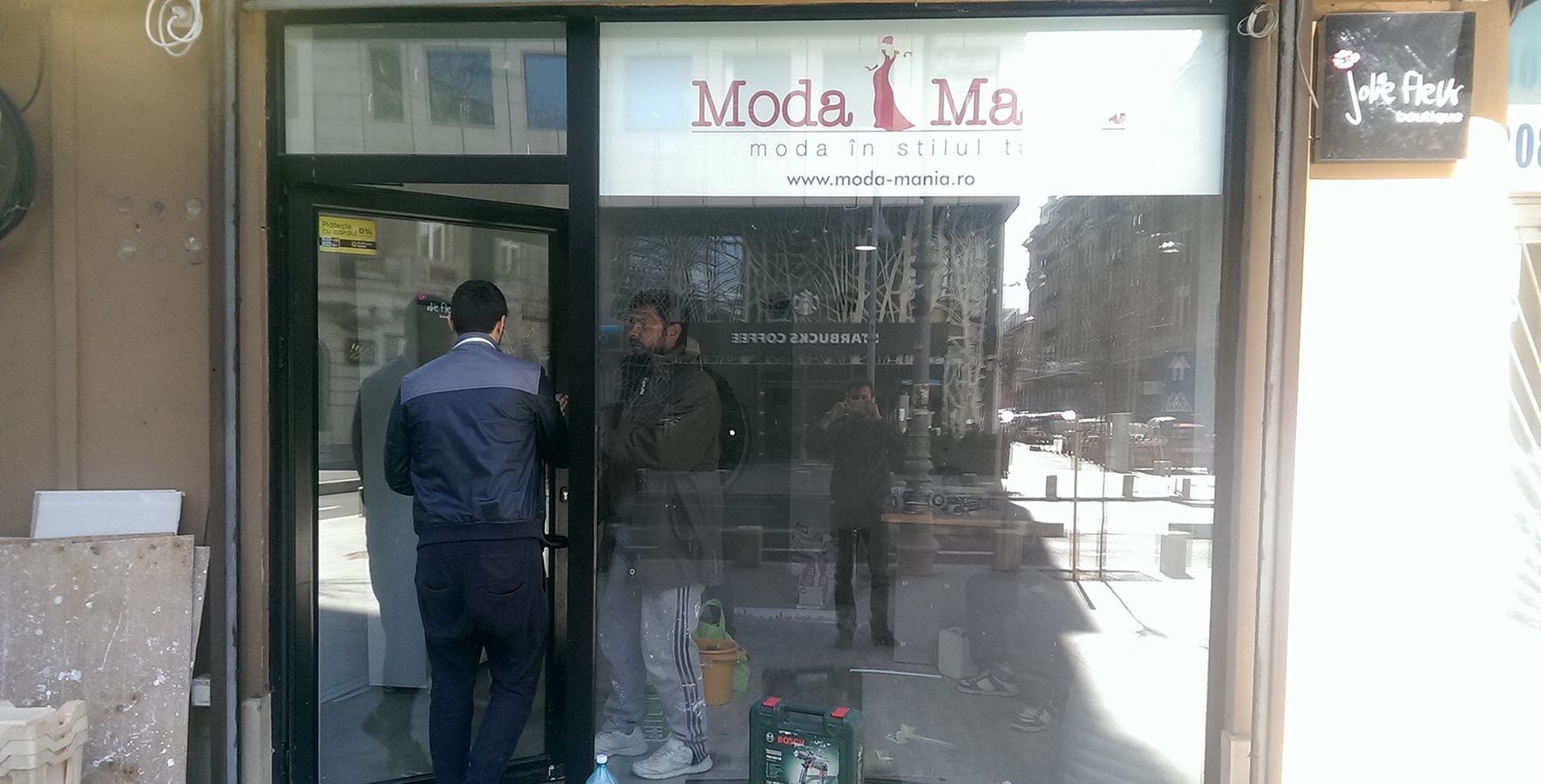 Ușa din aluminiu a magazinului ModaMania, deschisă, cu sculele lângă ea.